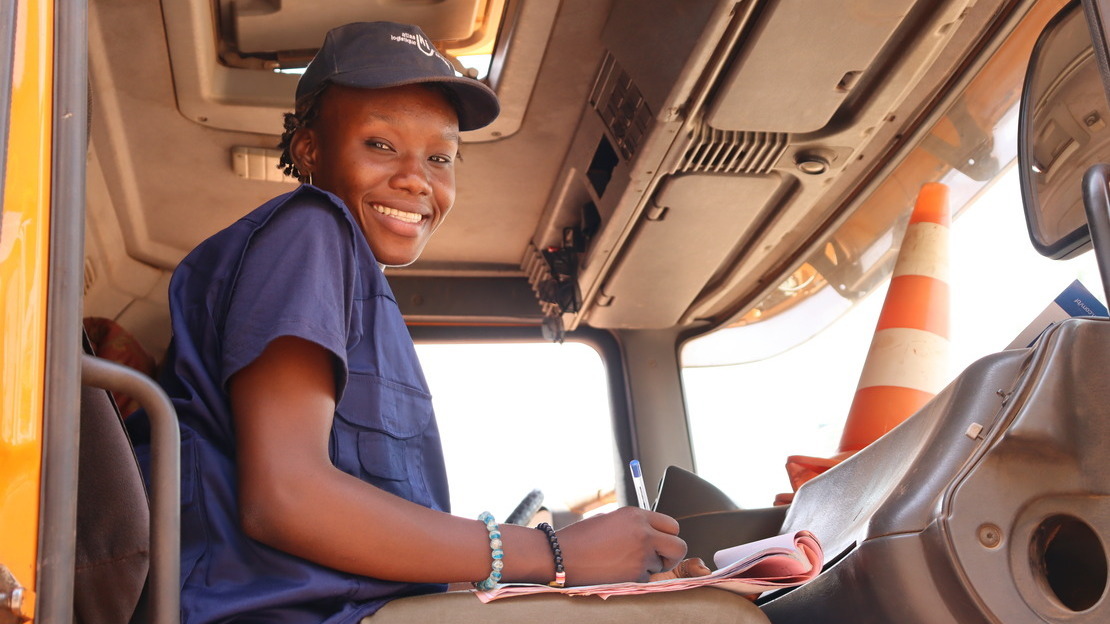 A young black woman sits in the cab of a truck. She's holding a pen and papers and looking down at the camera with a smile.; }}