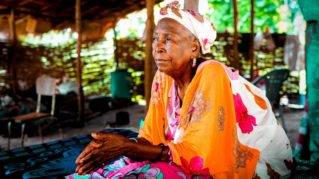 Portrait d'une femme âgée habillée de robe et de foulard colorés, assise à l'ombre d'une paillotte. Elle regarde vers la gauche et tient ses mains dans son giron.; }}