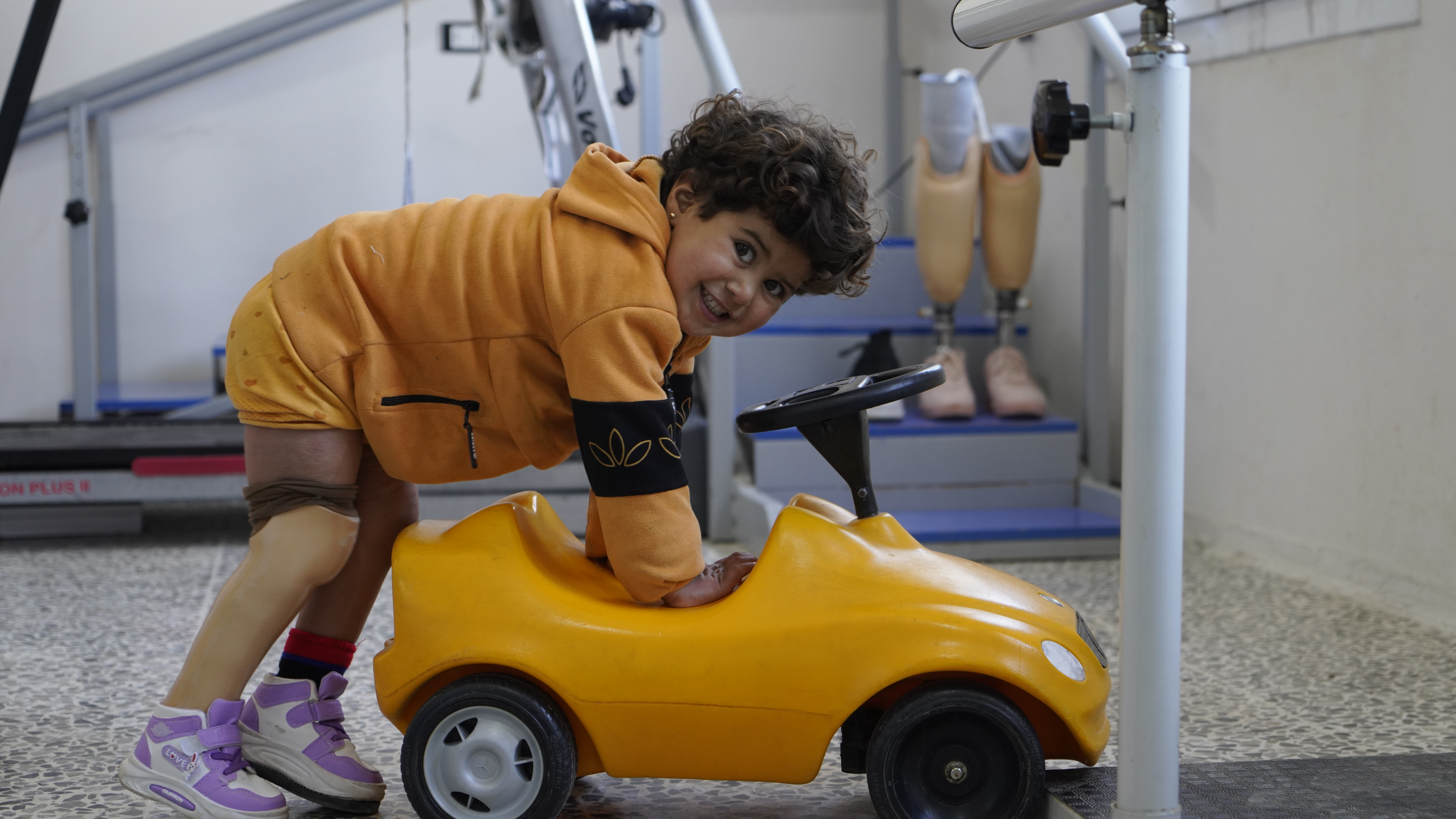 Noor during a rehabilitation session she jumps on a little yellow car smiling at the camera at Aqrabat hospital, HI’s partner in Northwest Syria. ; }}